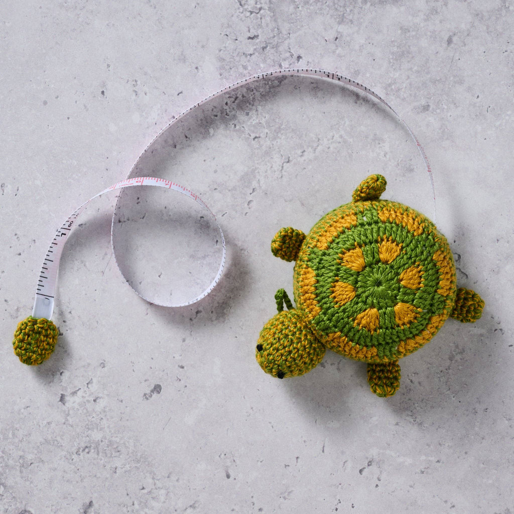 A measuring tape with a crocheted turtle design in green and dark yellow, displayed on a grey background.